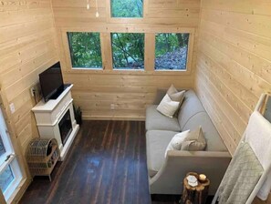A cozy cabin interior with a sofa, a fireplace, and three windows offering a view of the forest. - Mini Moonshine Ridge (Boone)