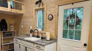 A compact kitchen with a sink, countertop, and cabinets. There is a window with a view of trees and a door with a peephole.