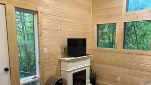 A cozy room with wooden walls, a fireplace, a television, and large windows offering a view of the forest.