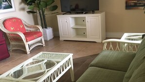 A living room with a green sofa, a white wicker coffee table, a wicker armchair, a white cabinet with a flat-screen TV, and a large potted plant. - 5 min walk to beach on inter coastal waterway (Fort Lauderdale)