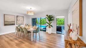 A spacious dining area with a wooden table set for six, surrounded by chairs with wooden legs. A large potted plant is in the center, and there are two framed artworks on the walls. - Chic + Upscale + Heated Pool | 15 min FLL & Beach (Plantation)