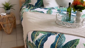 Una cama bien hecha con colcha de estampado tropical, almohada blanca texturizada y una cesta blanca decorativa al lado.