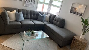 Una sala moderna con un sofá seccional gris, una mesa de centro de vidrio y una mesita auxiliar de mimbre con una planta.