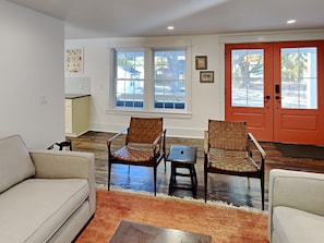 A living room with two wicker chairs, a small table, and a sofa. There are two windows with white frames and a red door.