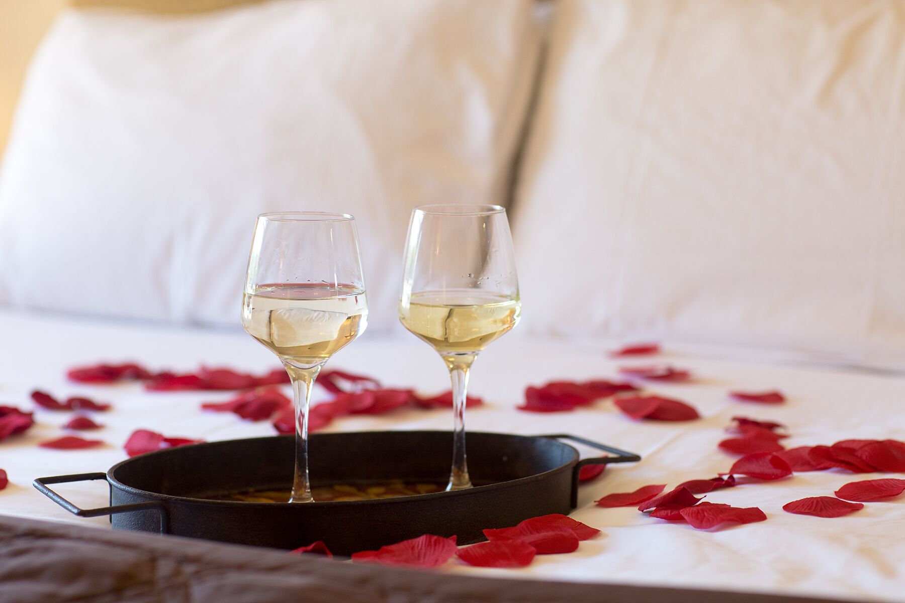 Two wine glasses on a tray with rose petals, placed on a bed with white linens.