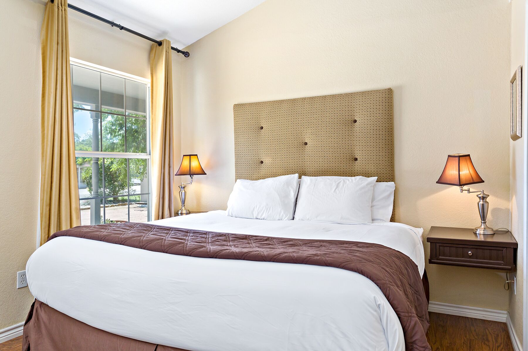 Quarto de hotel com uma cama grande, duas luminárias de cabeceira, cabeceira de vime e vista através de uma janela ampla.
