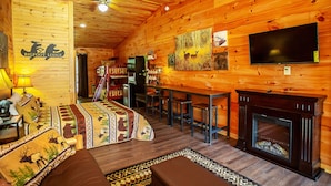 Interior de una cabaña de troncos con chimenea, un televisor montado en la pared y una zona de comedor con taburetes de bar.