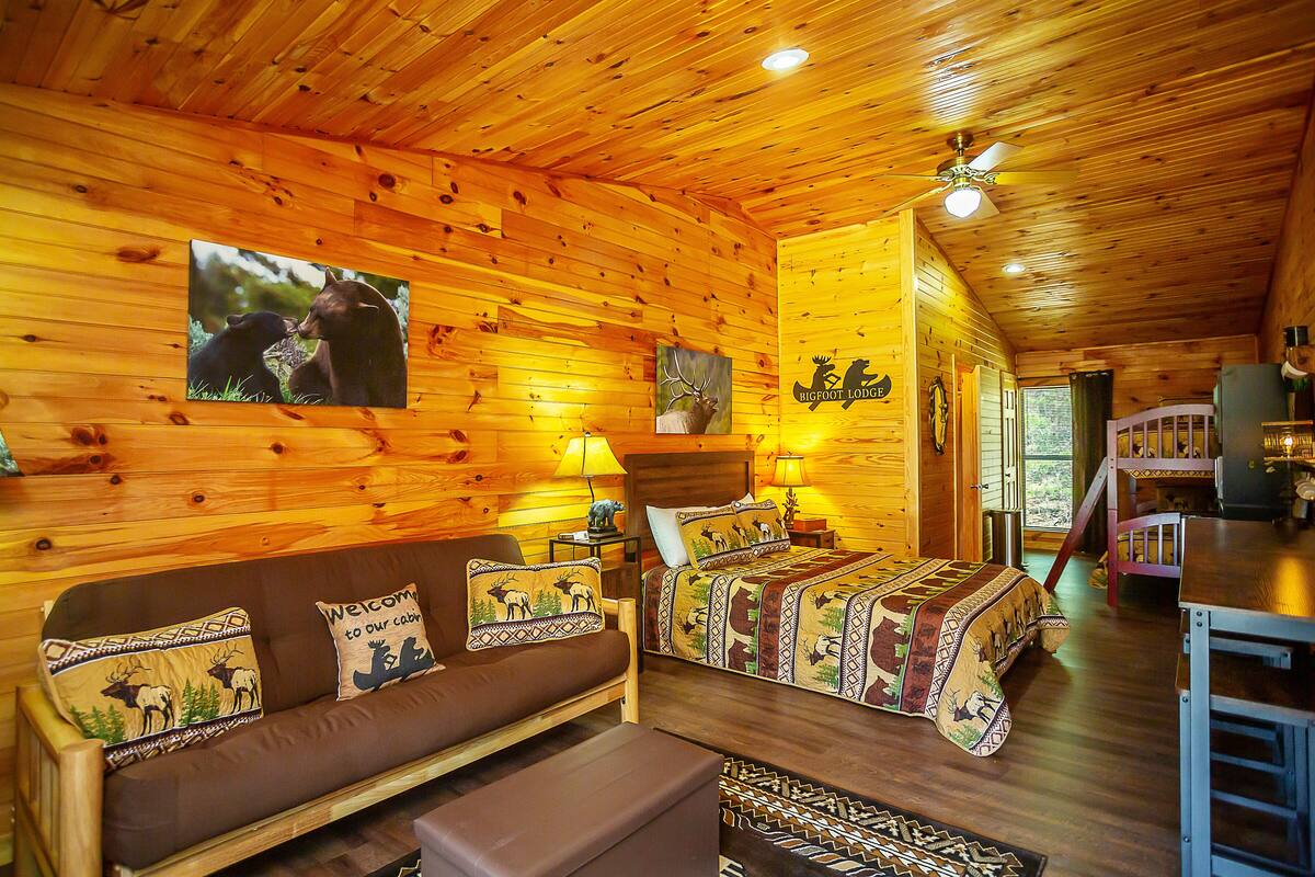 A cozy log cabin interior with a wooden ceiling, a sofa with animal-themed pillows, a bed with a patterned blanket, and a dining area.