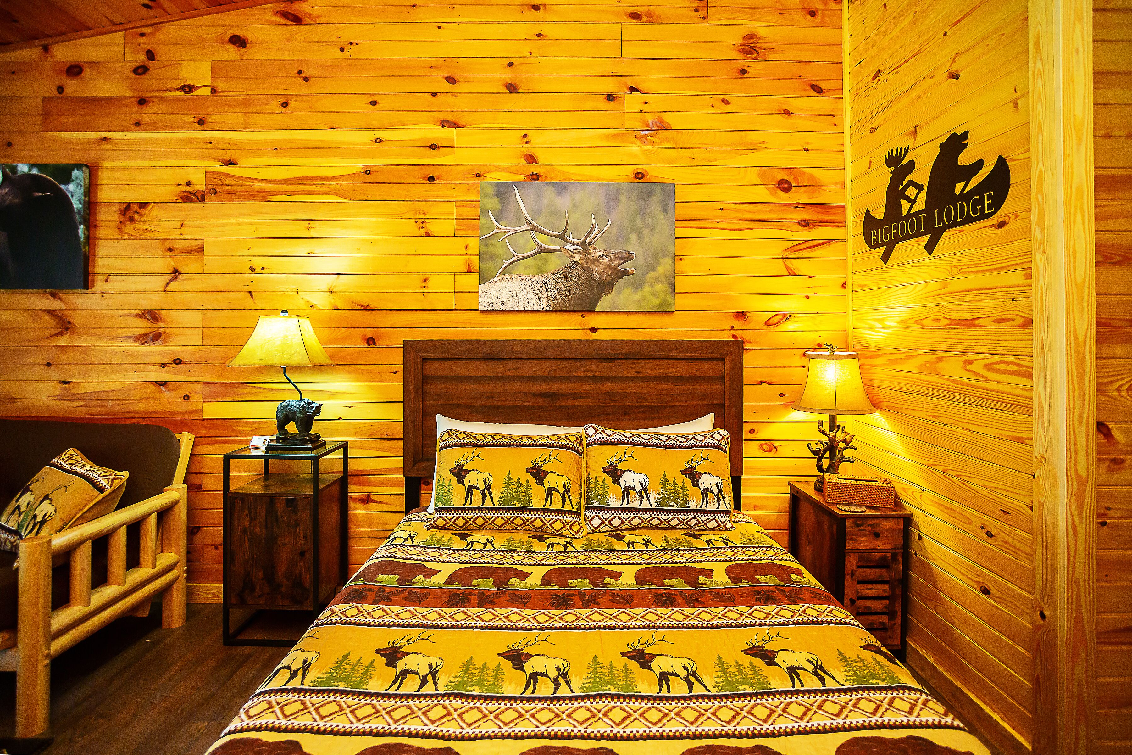 A log cabin-style room with a bed, two nightstands, a lamp, and a painting of an elk.