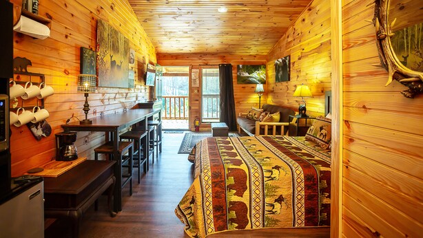 Interior acogedor de una cabaña con paredes de madera, un comedor, una cama con manta estampada y un espejo grande decorado con cuernos.