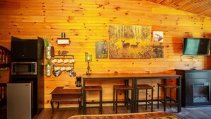 Interior acogedor de una cabaña con paredes de madera, una zona de comedor con mesa y taburetes, un refrigerador, un microondas y un televisor.