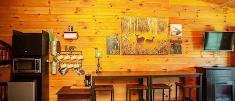A cozy cabin interior with wooden walls, a dining area with a table and stools, a refrigerator, a microwave, and a television.