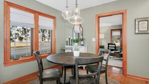 A dining area with a round table, four chairs, a chandelier, a window with blinds, and a mirror on the wall.