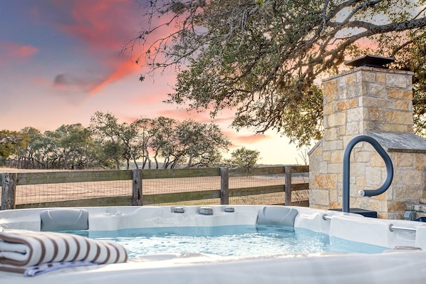 A hot tub with a stone chimney, surrounded by a wooden fence and trees, with a sunset in the background.