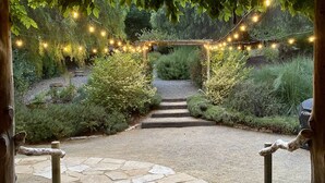 Un sendero de piedra que conduce a una pérgola de madera decorada con luces de cadena, rodeada de abundante vegetación.