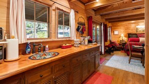 Interior de una cabaña de madera con área de cocina, que incluye encimera de madera, lavamanos y ventana con vistas a un jardín.