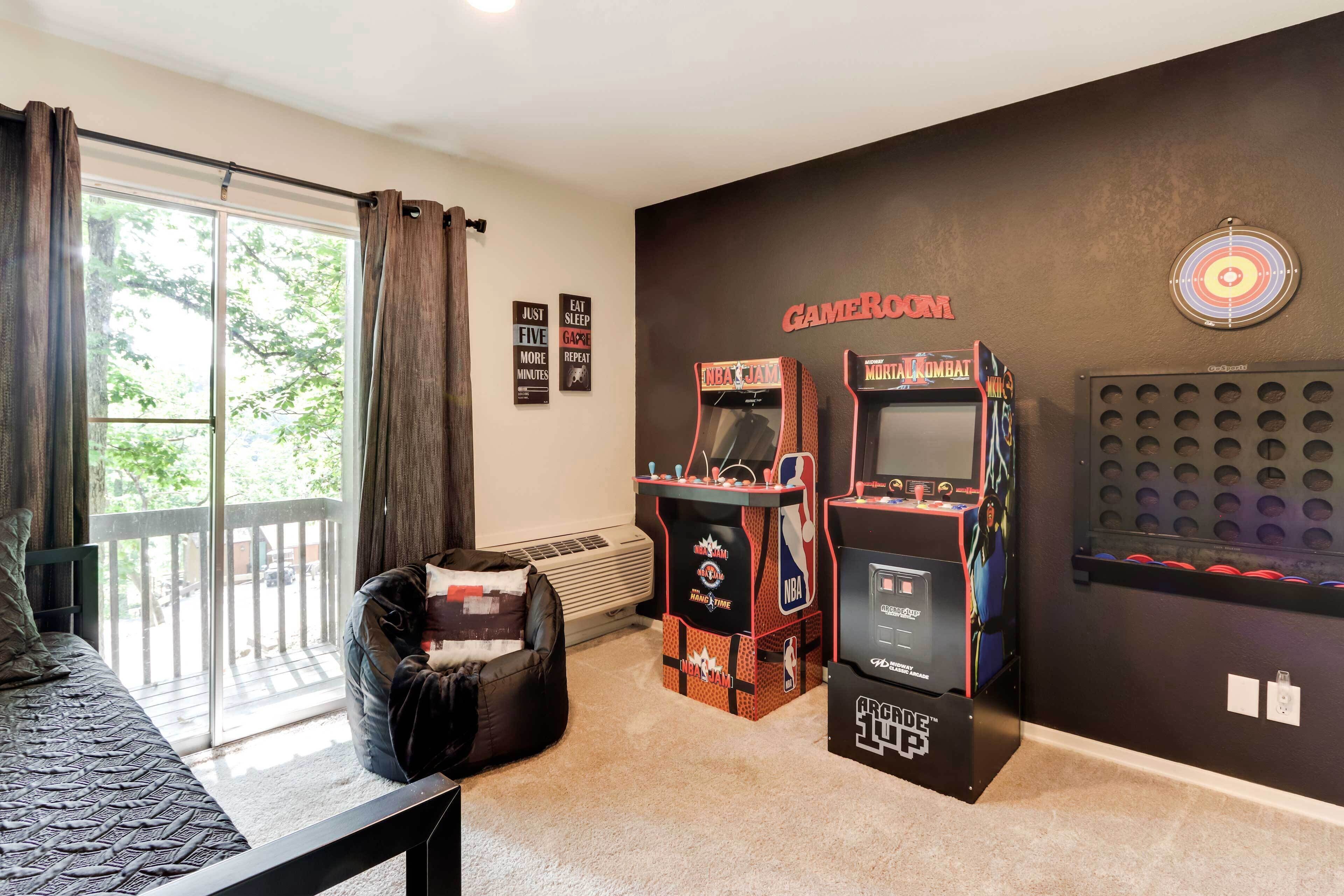 A room with two arcade machines, a bean bag chair, a bed, and a large window.