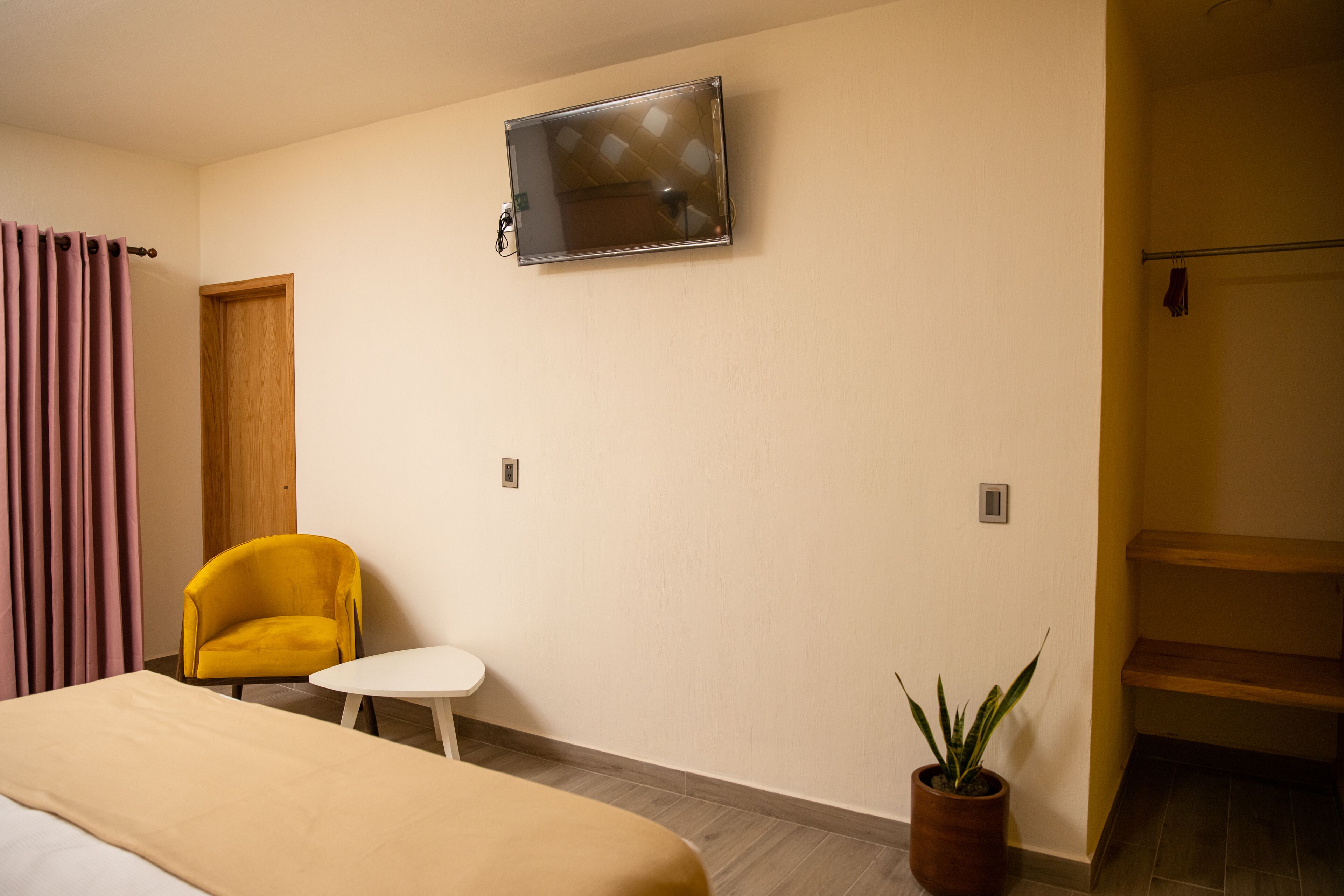 Habitación de hotel con cama, una pequeña mesa blanca, un sillón amarillo, una planta en maceta, un televisor montado en la pared y una puerta de madera.