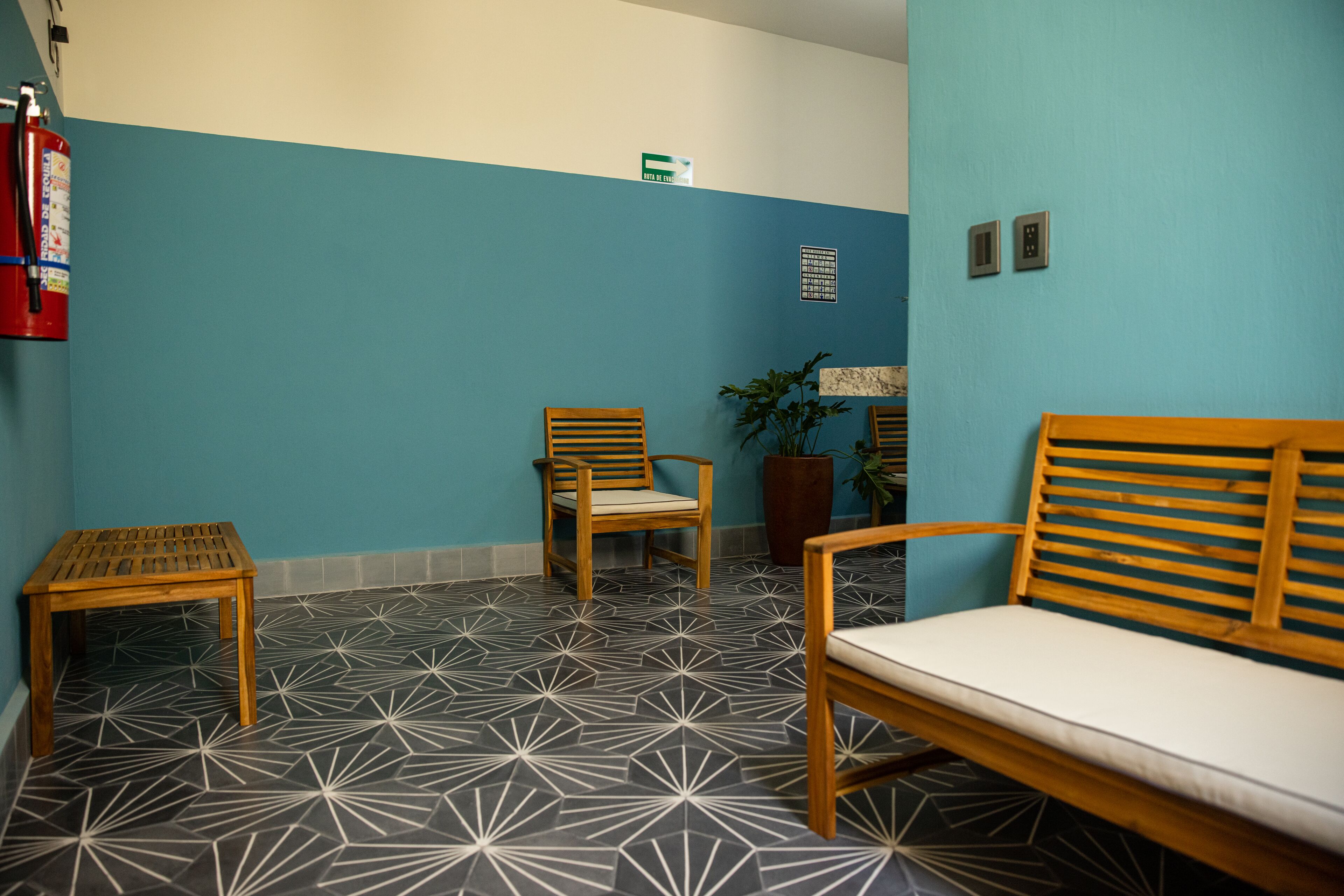 Un área de espera con pared azul, un extintor, dos sillas de madera, una mesita y una planta en maceta.