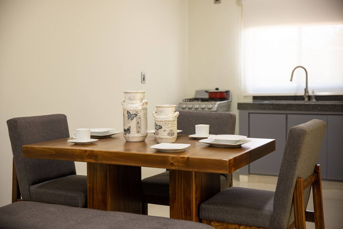 A dining table with chairs, a kitchen in the background, and a window with a sink.