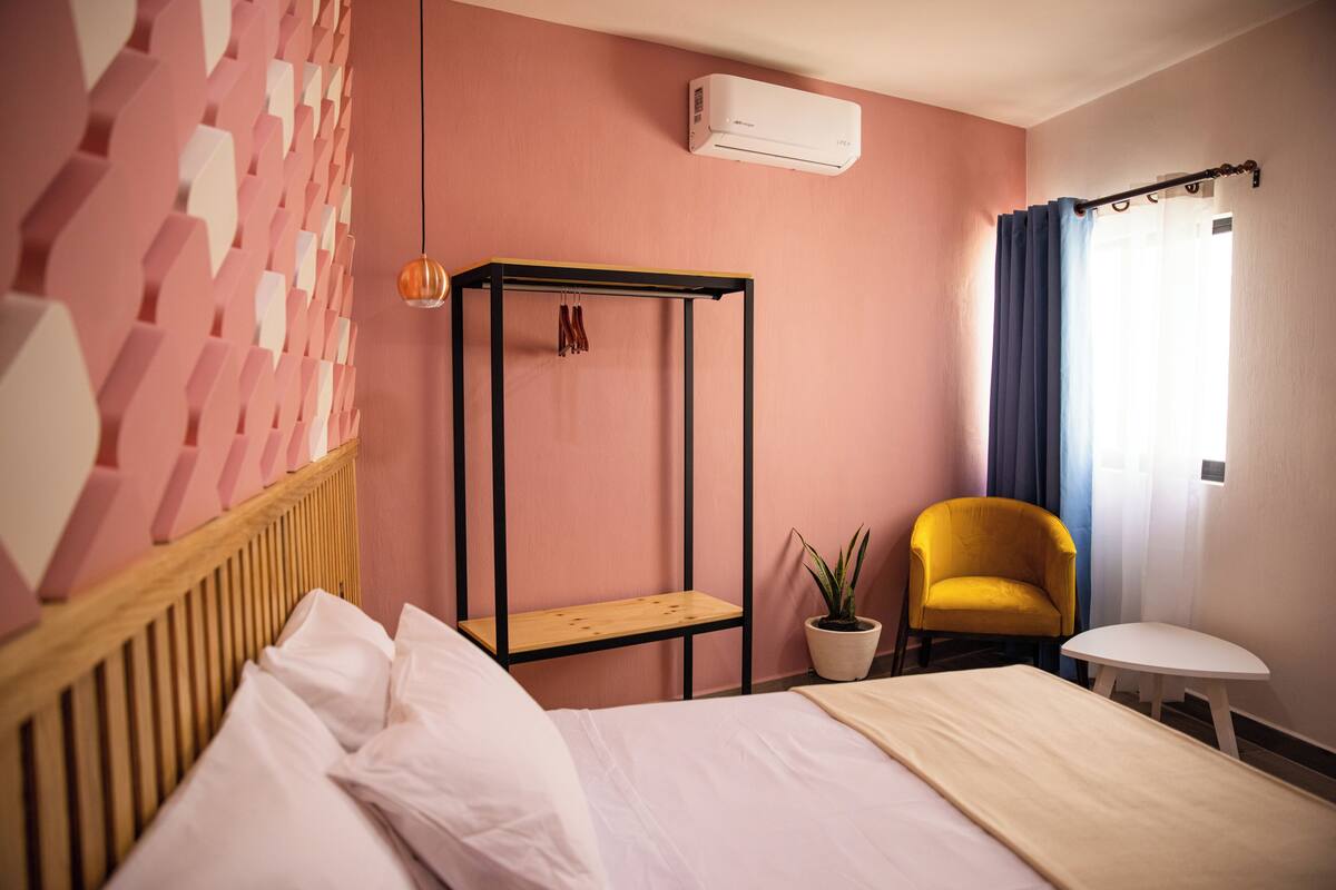 A bedroom with a bed, a wooden headboard, a wall-mounted air conditioner, a small table, a chair, and a potted plant.