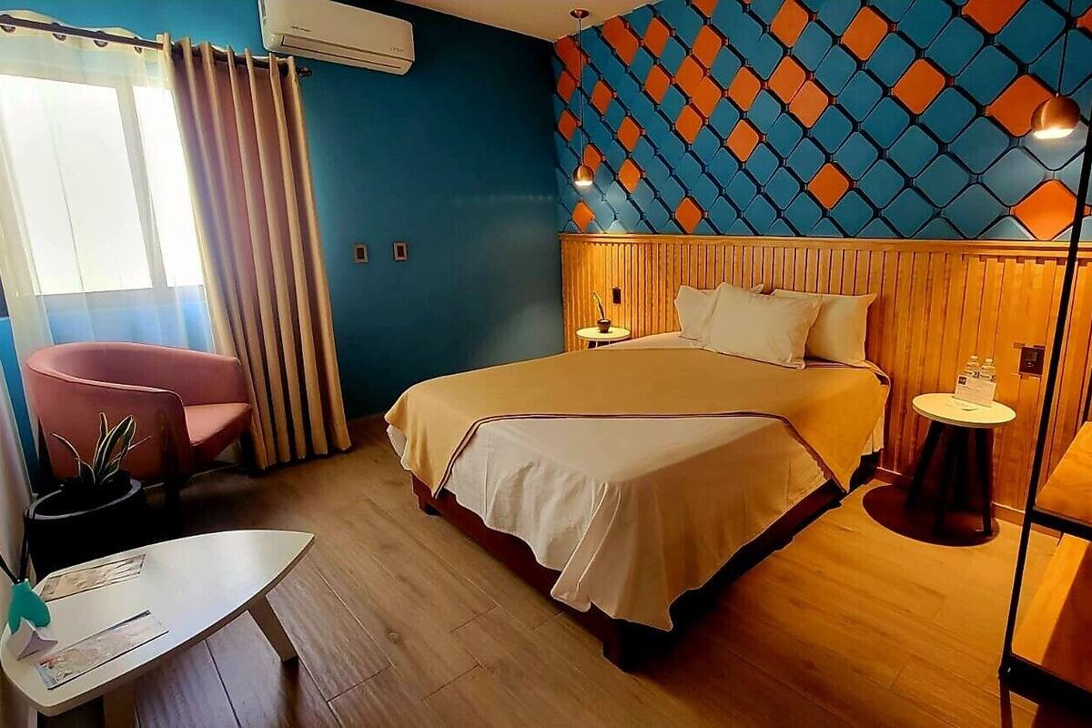 A hotel room with a bed, a small table, a chair, and a wall with a geometric pattern.