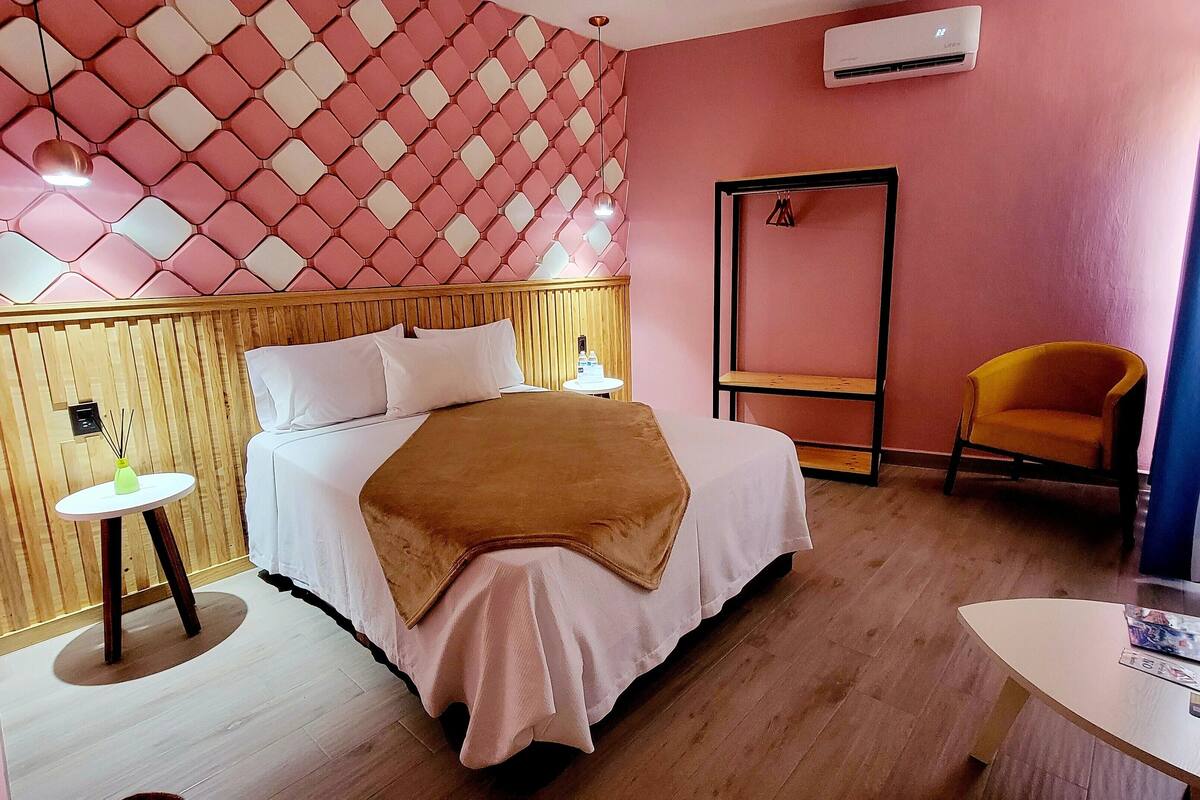 A hotel room with a bed, bedside table, chair, and a wall-mounted air conditioner.