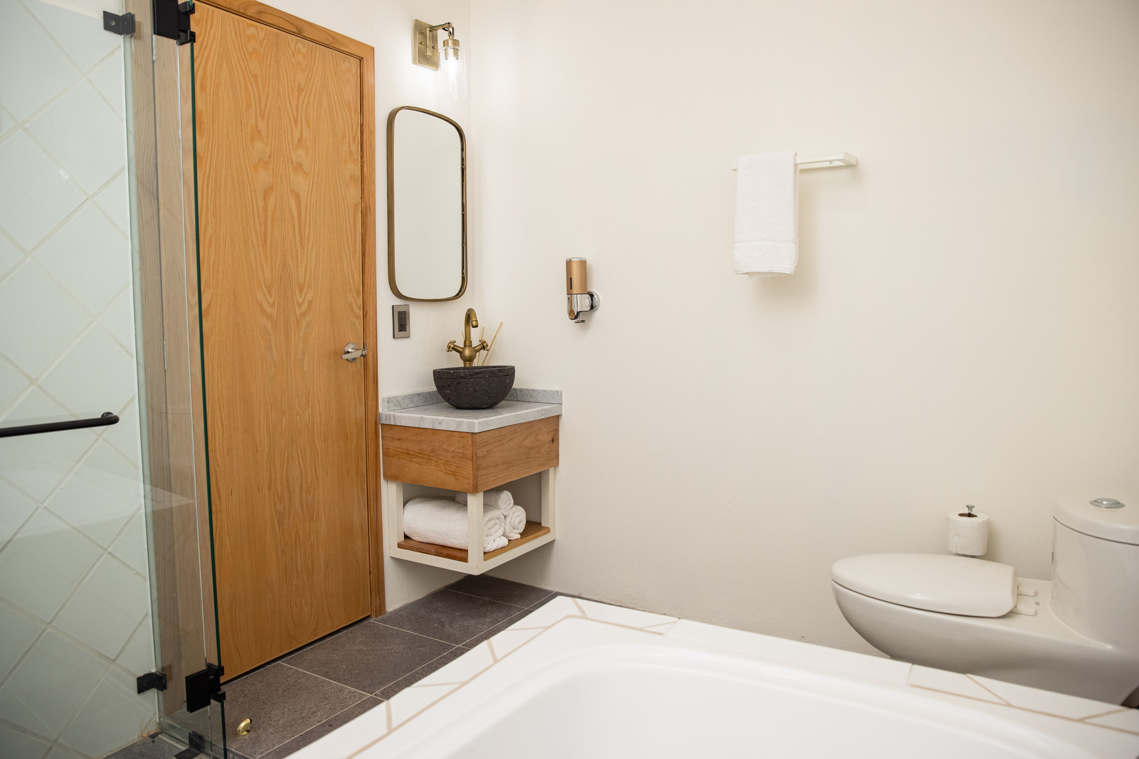 A bathroom with a glass shower enclosure, a wooden door, a sink with a marble countertop, a toilet, and a white bathtub.