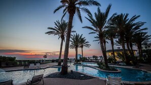 A pool area with palm trees and lounge chairs, surrounded by a building with a view of the sunset. - STUNNING BEACHFRONT WITH TRIPLE WATER VIEWS OF OCEAN, LAKE, BAY! (Galveston)