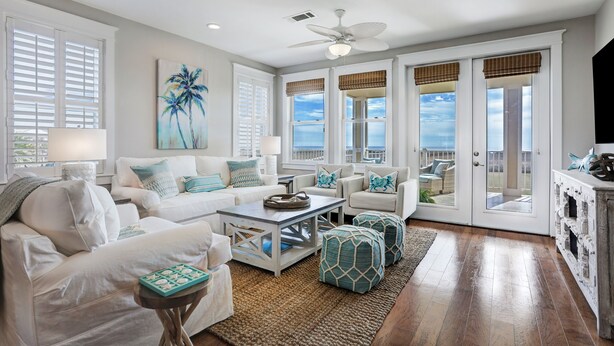 A spacious living room with white furniture, a ceiling fan, and large windows with views of the ocean. - STUNNING BEACHFRONT WITH TRIPLE WATER VIEWS OF OCEAN, LAKE, BAY! (Galveston)
