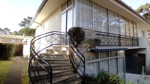 Una casa de dos pisos con una escalera que lleva a la entrada, ventanas amplias y un pequeño jardín.