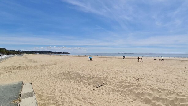 Una amplia playa de arena con cielo despejado, algunas personas y una costa distante.