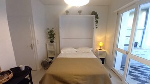 Un dormitorio con cama, mesitas de noche, un escritorio y una silla. Hay plantas en las mesitas de noche y una ventana con vistas al exterior.
