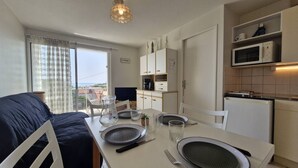 A compact room with a dining table set for two, a kitchenette with a microwave and refrigerator, and a window with curtains. - Royan - APARTMENT with SEA VIEW - ONLY 300m from the BEACH and SHOPS (ROYAN)
