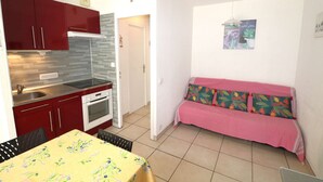A compact kitchen with red cabinets, a dining table with a floral tablecloth, and a pink sofa with patterned cushions.