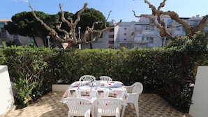 A patio with a table set for four, surrounded by white chairs, with a hedge and buildings in the background.