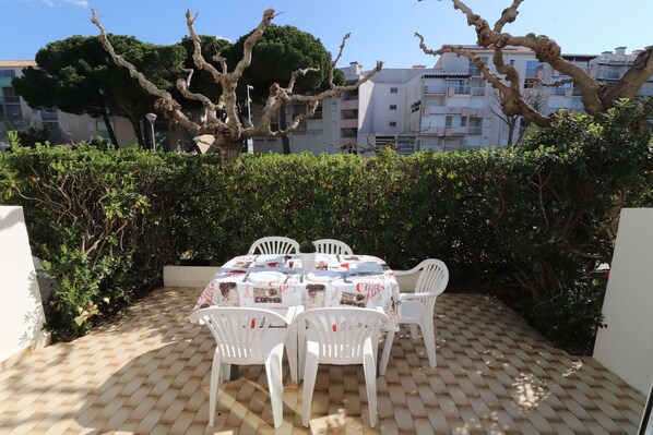 A patio with a table set for four, surrounded by white chairs, with a hedge and buildings in the background.