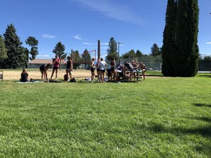 Un groupe de personnes s’est rassemblé autour d’un filet de volley-ball dans un champ herbeux, avec des arbres et une clôture en arrière-plan.