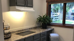 A kitchen with grey cabinets, a granite countertop, a black induction cooktop, a potted plant, and a window with a view of a backyard. - Cozy Studio (Sandpoint)