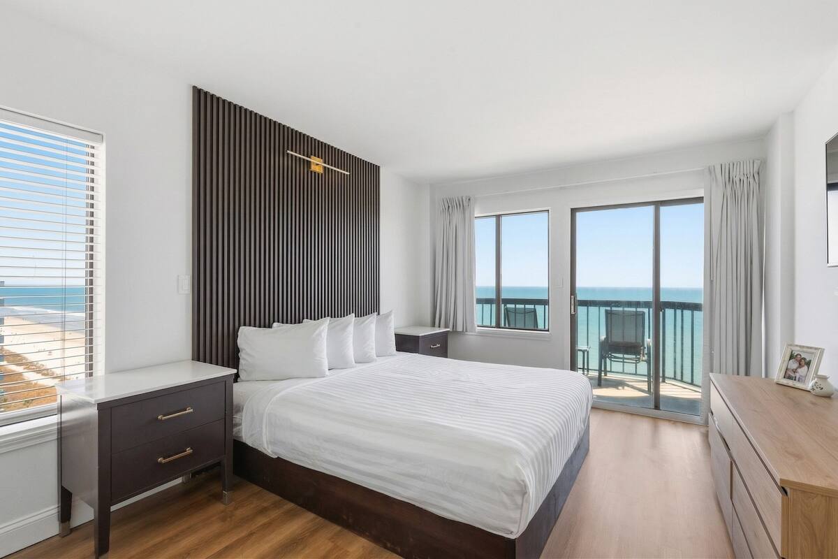Un dormitorio con una cama grande, una mesita de noche, un aparador y un balcón con vistas al mar.