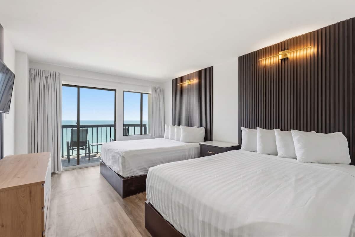 Habitación de hotel con dos camas, un escritorio de madera, un balcón con vista al mar y un diseño moderno en la pared.