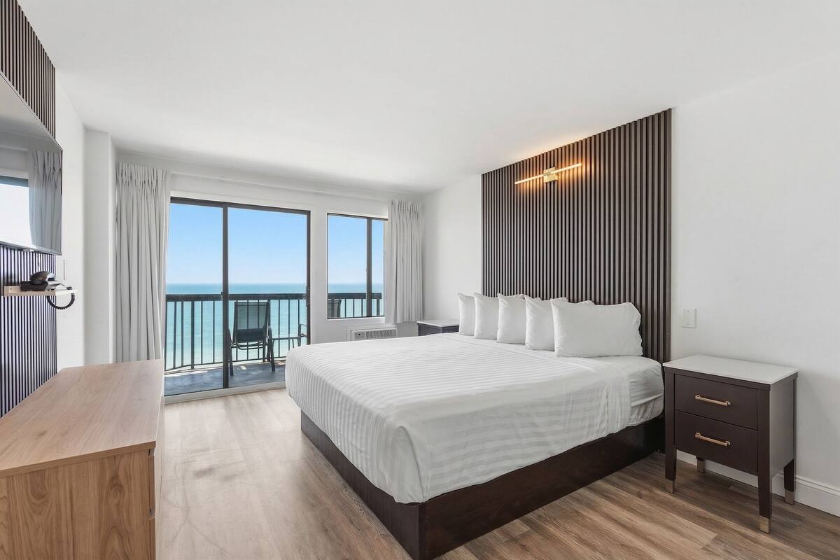 Una habitación de hotel moderna con una cama grande, una mesita de noche, un escritorio de madera y un balcón con vistas al mar.