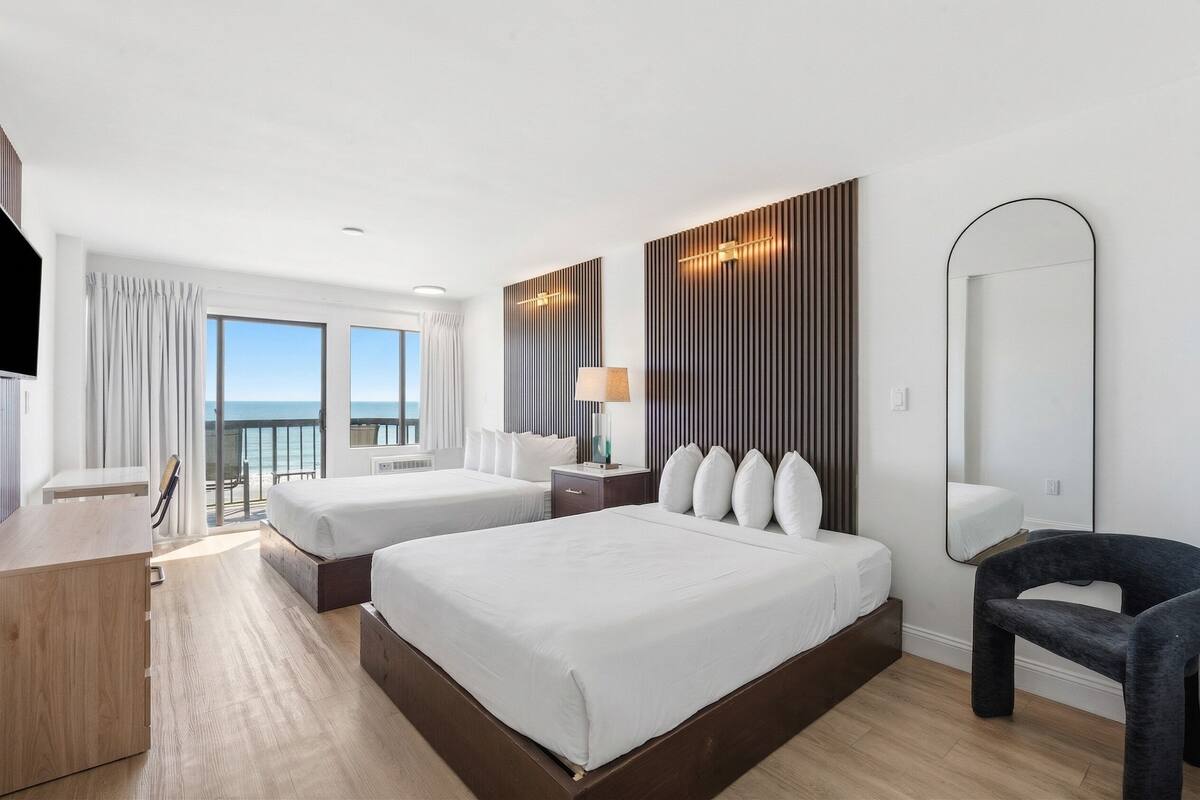 Habitación de hotel con dos camas, un escritorio, una silla, un espejo grande y vista al océano.