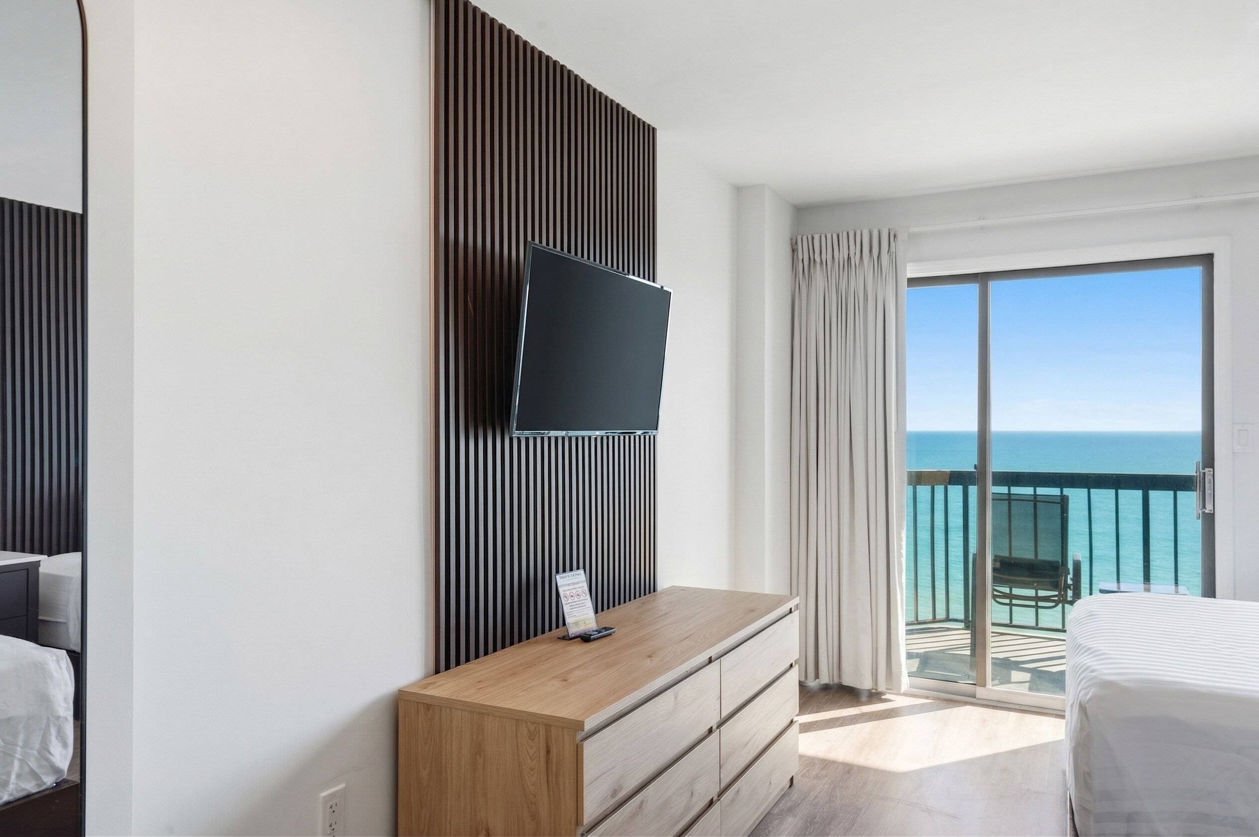 A hotel room with a TV mounted on a wall, a wooden dresser, a bed, and a balcony with a view of the ocean.