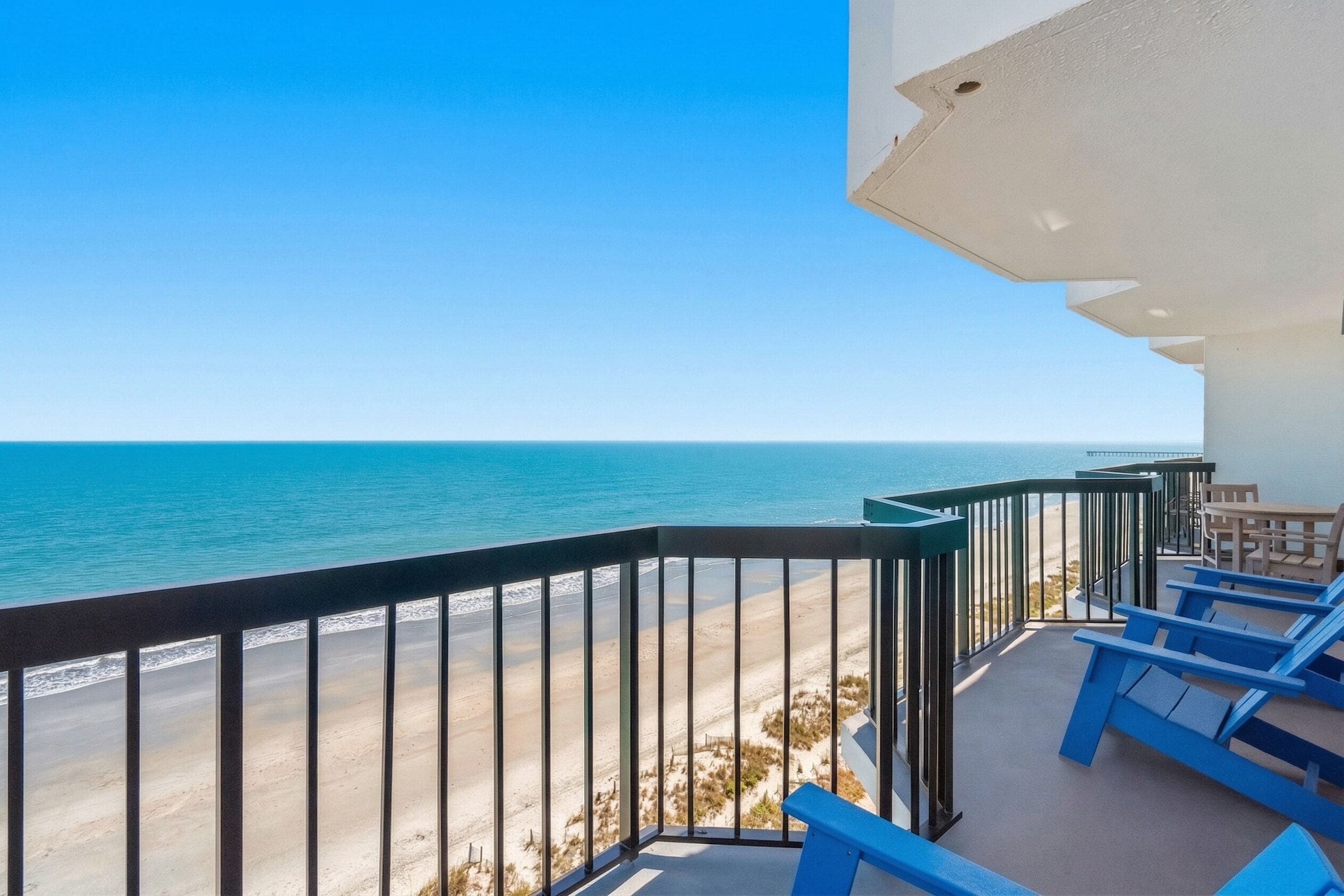 A balcony with a view of the beach and ocean, blue lounge chairs, and a table.