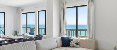 A modern living room with a large sectional sofa, a dining table with chairs, and large windows offering a view of the sea.