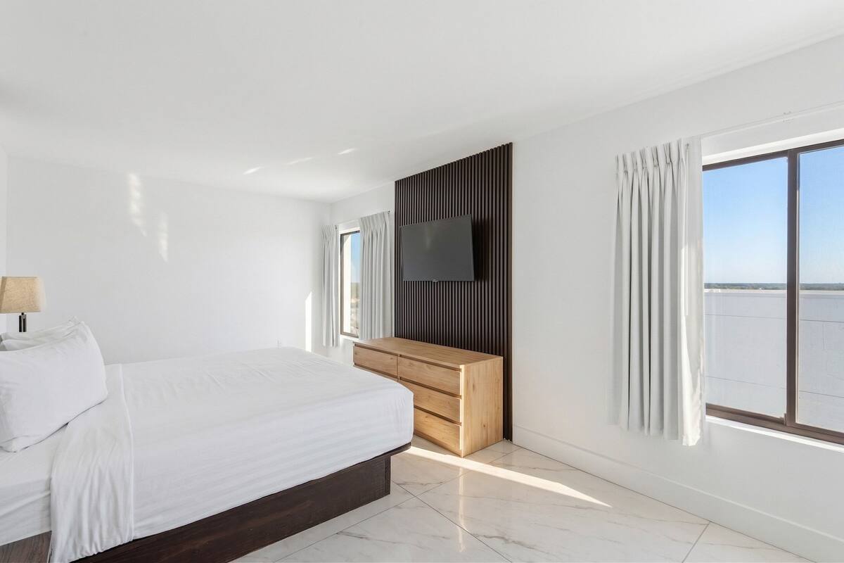 Una habitación de hotel moderna con una cama grande, un aparador de madera, un televisor de pantalla plana y un ventanal con vistas al mar.