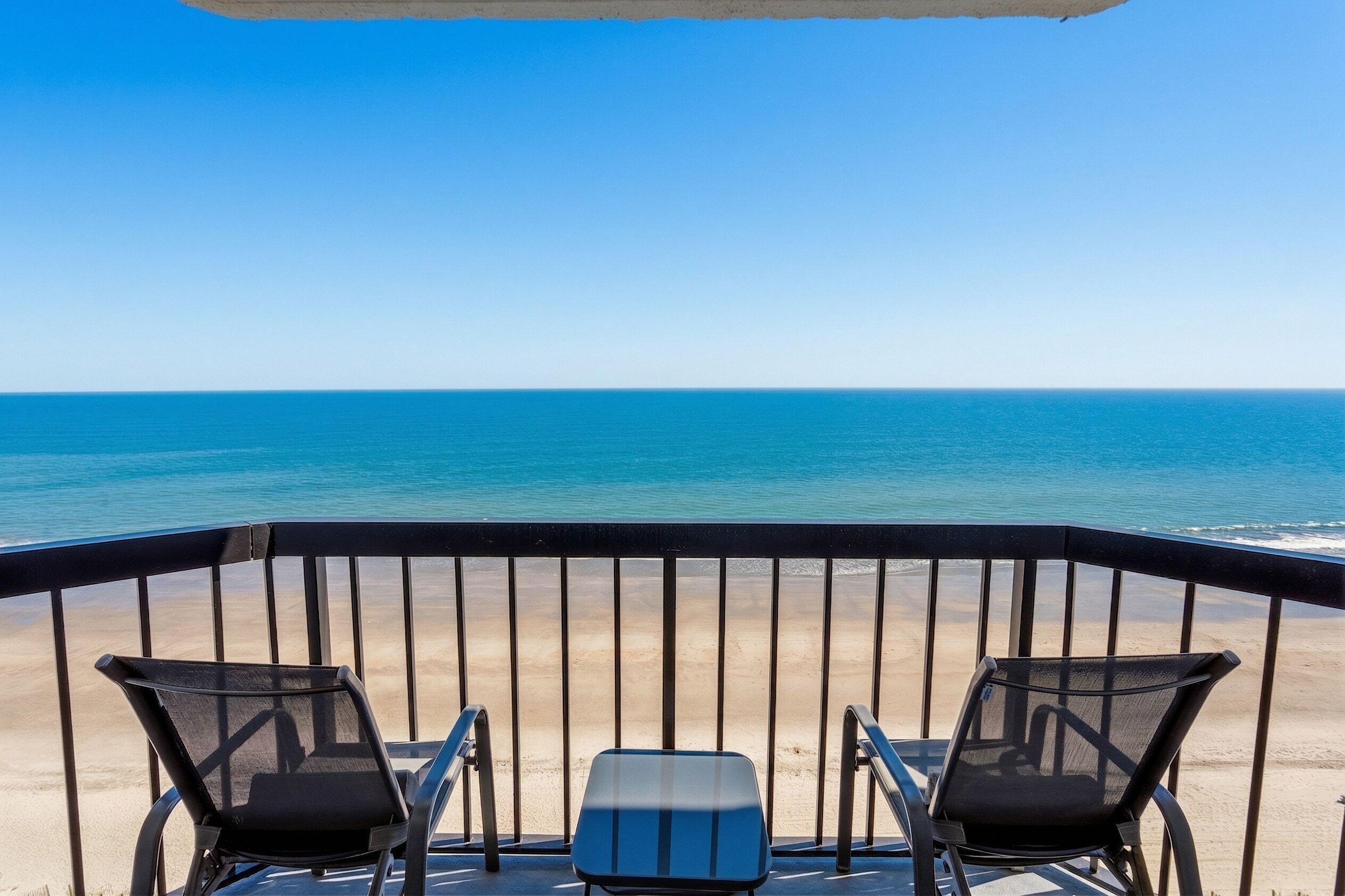 Ein Balkon mit zwei Stühlen und einem kleinen Tisch, der Blick auf einen Strand und den Ozean gewährt.
