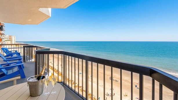 A balcony with a view of the beach, blue chairs, and a table with a champagne bucket. - Grande Beach Resort (Myrtle Beach)