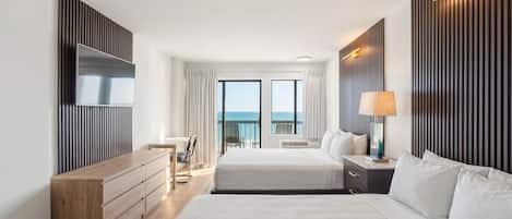 A hotel room with a large bed, a desk, a chair, a TV, and a view of the sea.